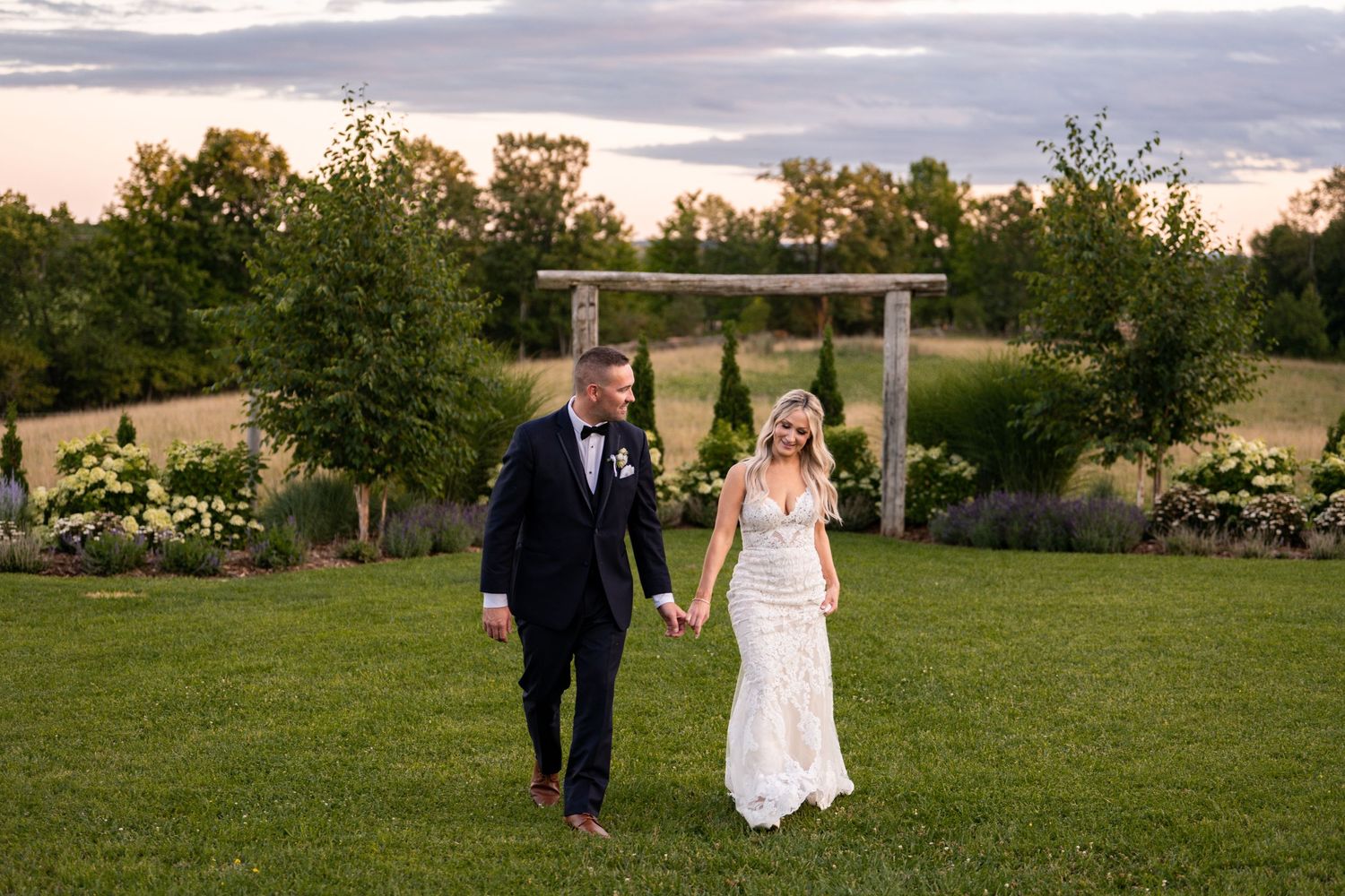 Couple walking together on grass with wooden arbor and sunset sky in background.