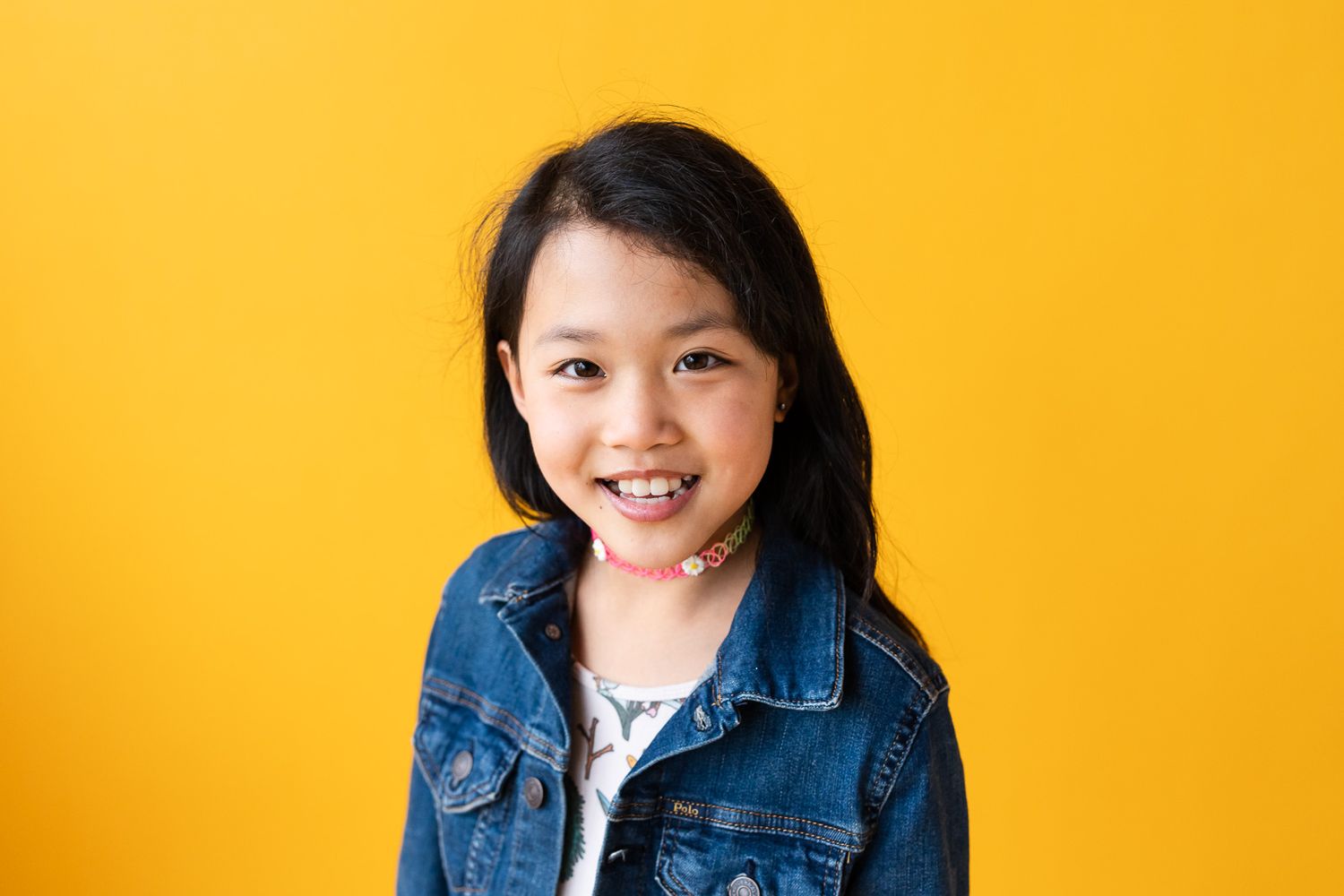 Smiling portrait of a girl wearing a jean jacket and dress in front of a yellow backdrop