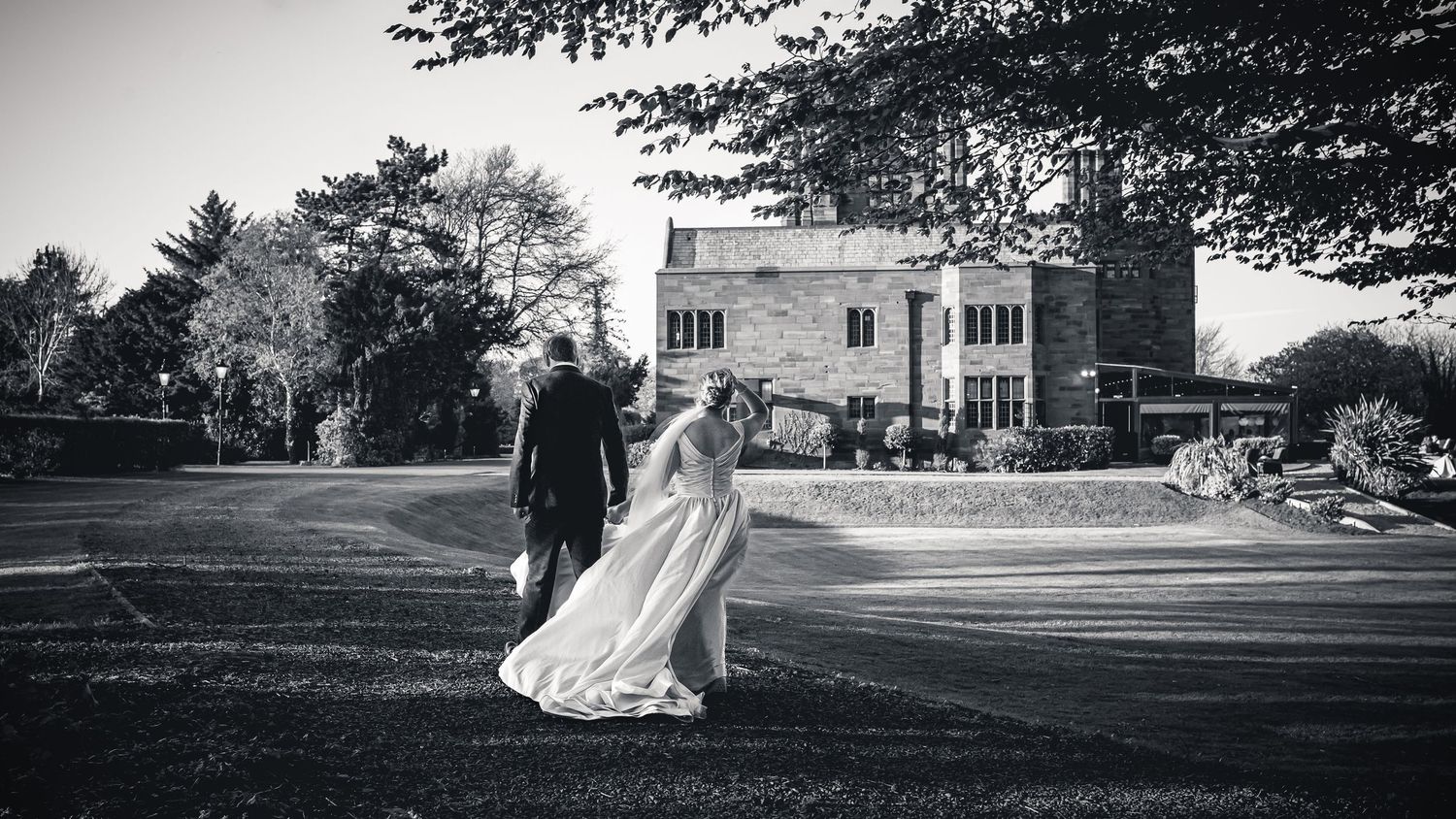 A black and white photo of newlyweds walking across a grand estate lawn.