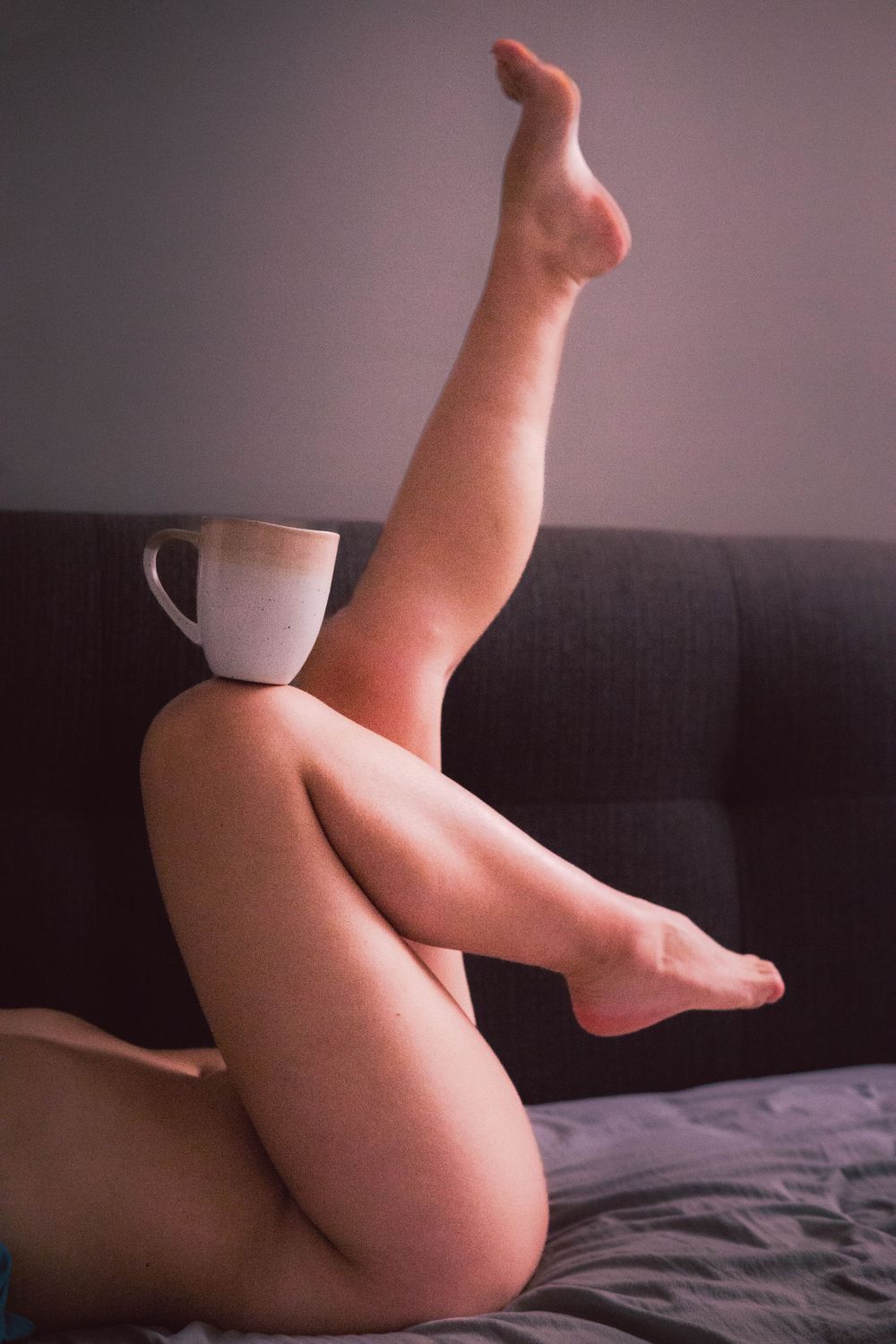 Artistic photo of bare legs propped up on a dark couch with a white coffee mug balanced on the knee.