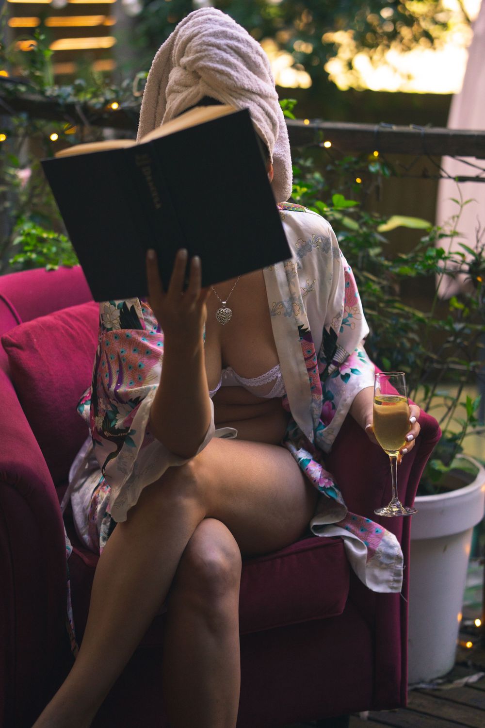 Person wearing underwear in floral robe reading book with champagne on red velvet chair in garden setting.