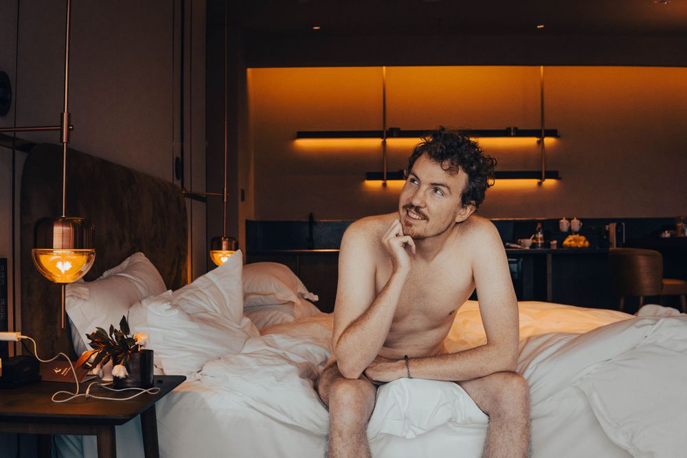 Nude person sitting on edge of bed in dimly lit hotel room with warm ambient lighting.