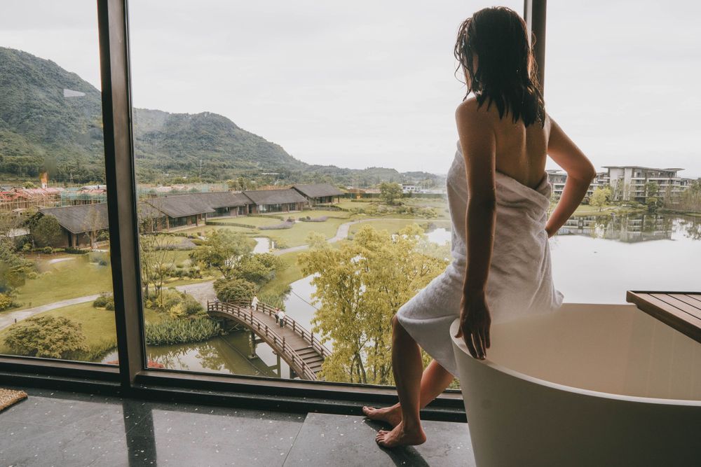 Figure in white towel gazes out large window overlooking mountainous landscape and luxury bathtub.