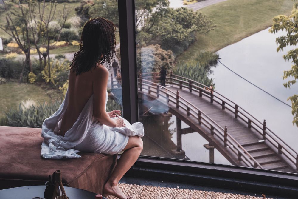 Person in a white dress sits at a window overlooking a curved wooden bridge and tranquil lake view.