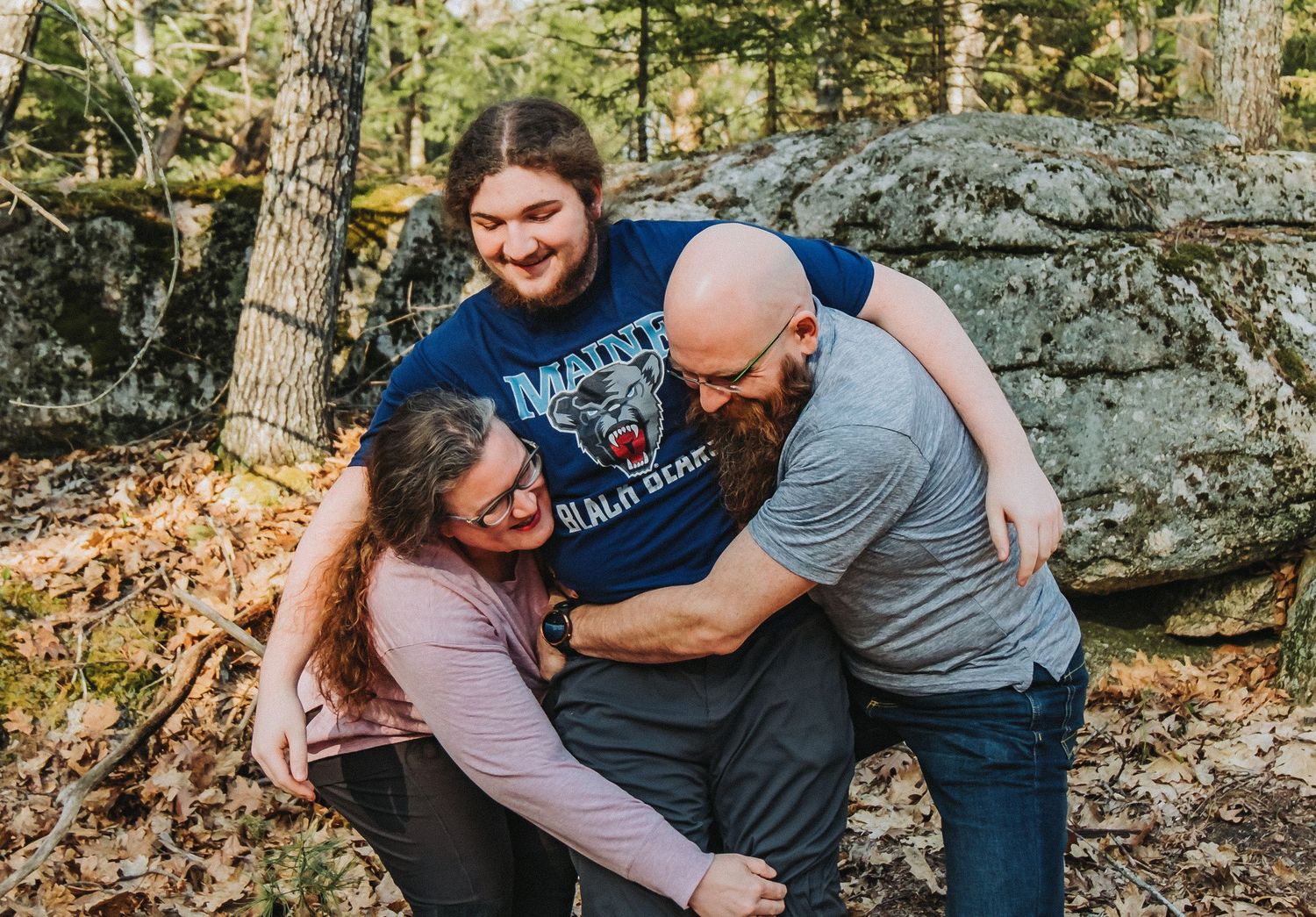 A mom, dad, and their son who is a senior share an embrace while enjoying the outdoors among large boulders and trees in a forest setting.
