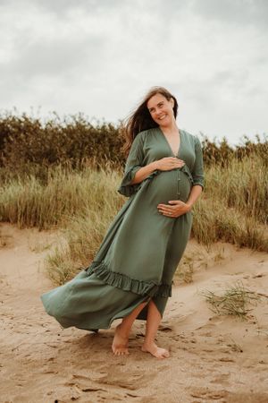 Woman in olive green dress poses gracefully on sandy beach during sunset maternity session.