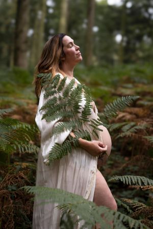 Woman in white dress holding fern branches in a forest setting with dreamy natural lighting.