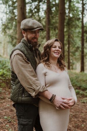 An expectant couple embraces tenderly in a peaceful forest setting.