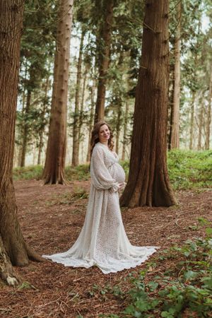 A white lace maternity gown creates elegant silhouettes against towering forest trees.