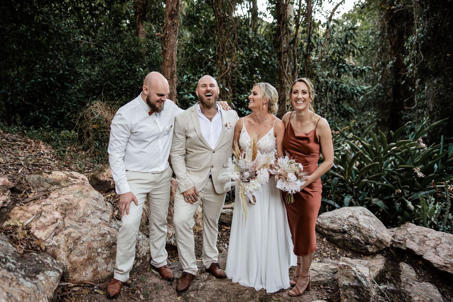 Wedding party in light colored attire poses together on large rocks surrounded by lush greenery.
