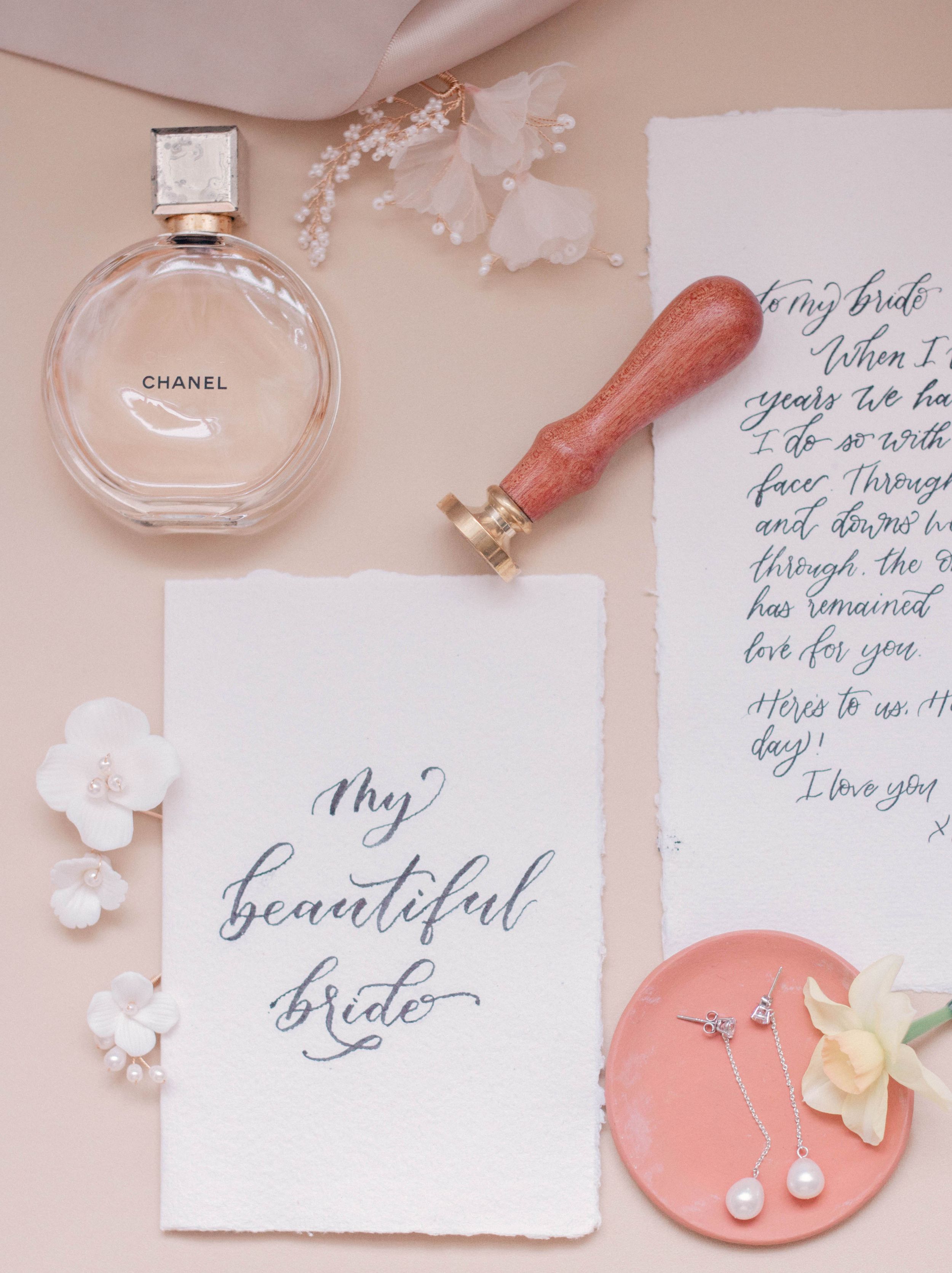 Wedding details flatlay with calligraphy notes, perfume bottle, and coral accents.