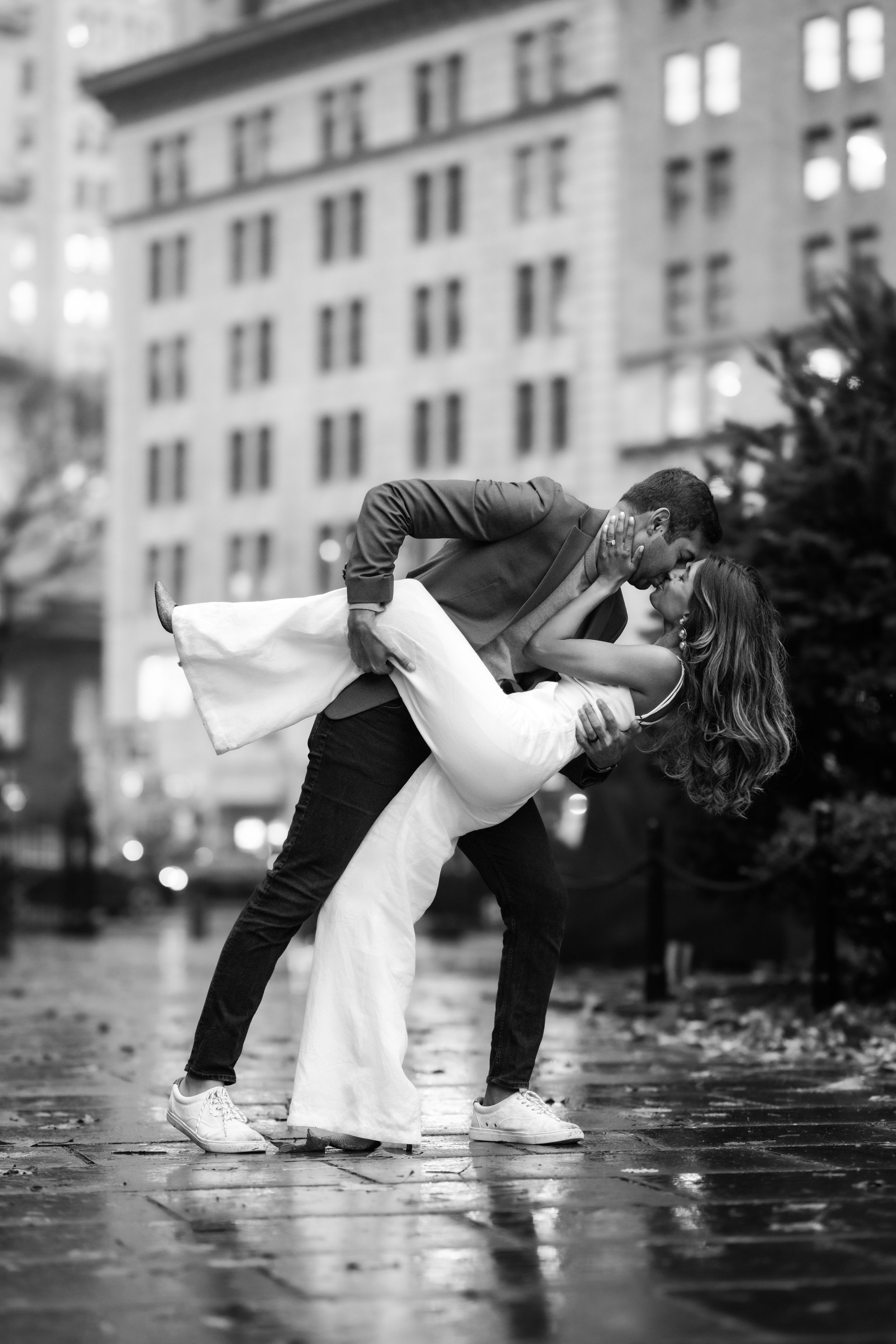 Black and white photo of man dipping woman in white dress on rainy city street at night.