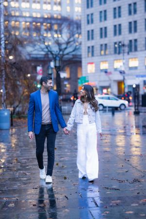 Couple walking hand in hand on rain-soaked city plaza, woman in white and man in blue blazer.