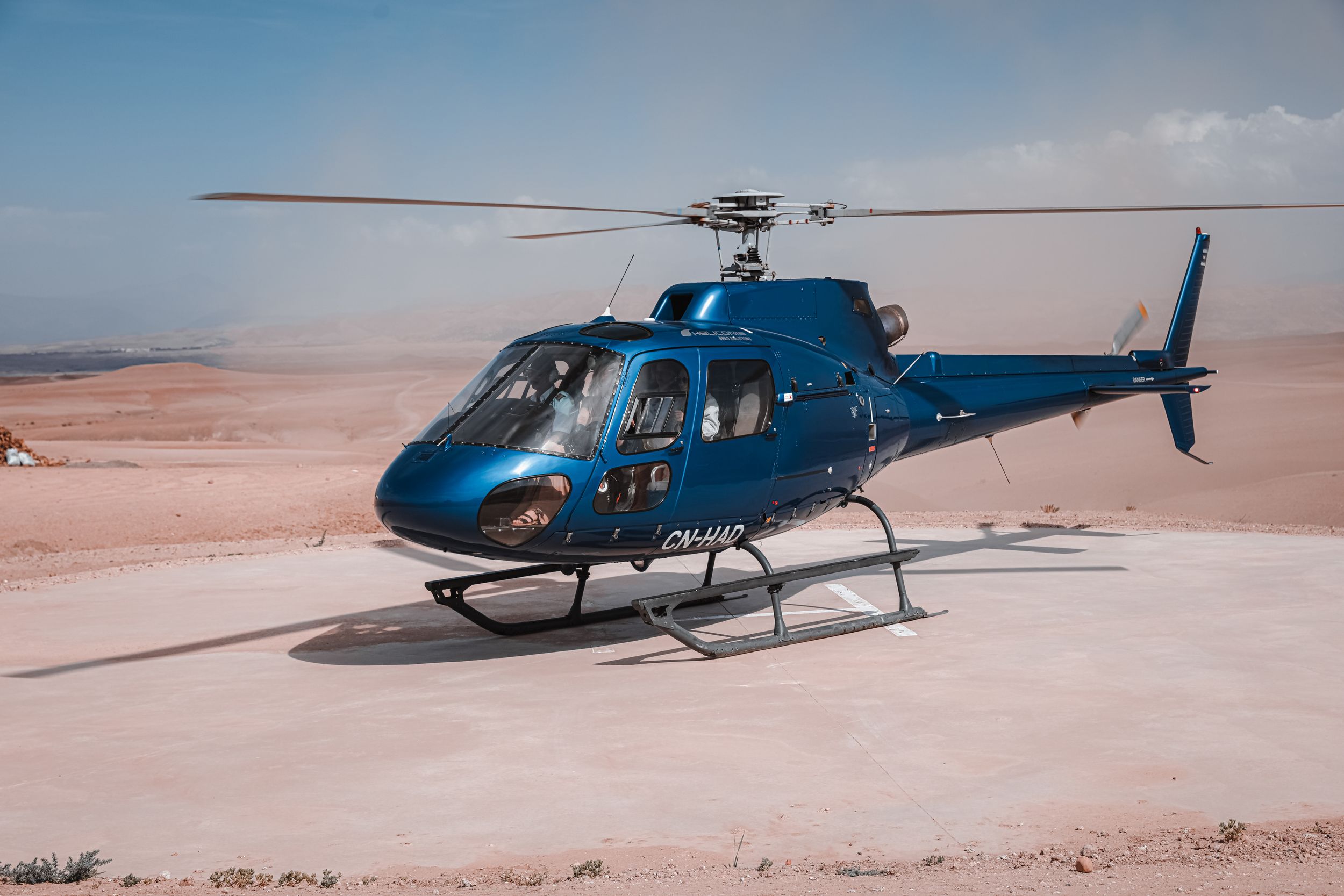 A blue helicopter sits on sandy terrain against a bright sky during daytime.