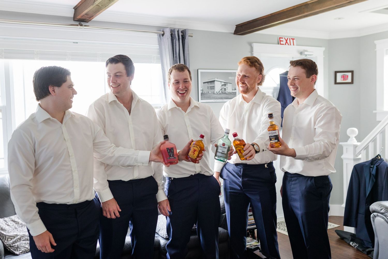 Groomsmen in white shirts and navy suits share laughter while getting ready on wedding day.