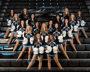 High school dance team posed in formation on bleachers wearing blue and white uniforms with pom poms at a sports venue.
