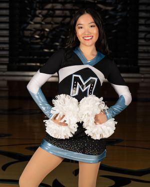 A high school dance team dancer in a black and blue uniform with white pom poms poses in a gymnasium.