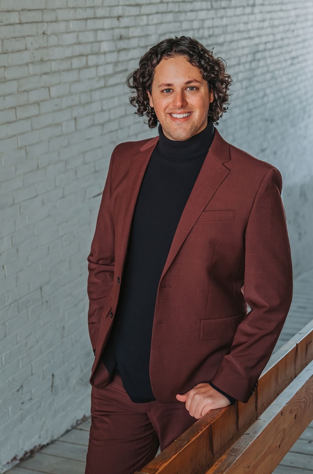 Professional headshot of someone wearing a burgundy suit jacket and black turtleneck against a white brick wall. Taken in Bath, Maine.