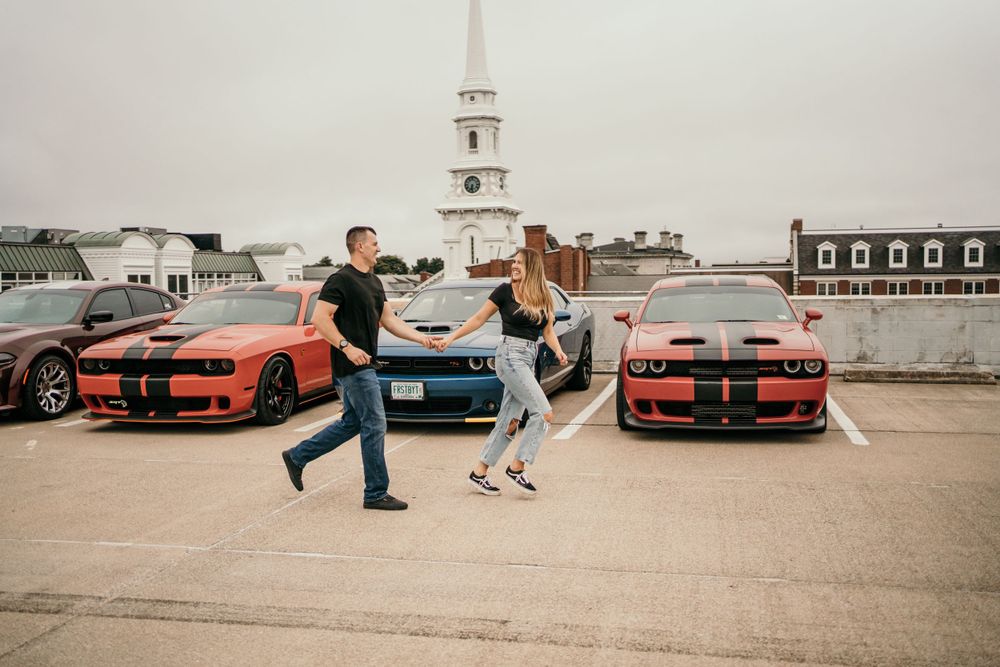 Playful couple dancing in parking lot with vintage cars and church spire in background.