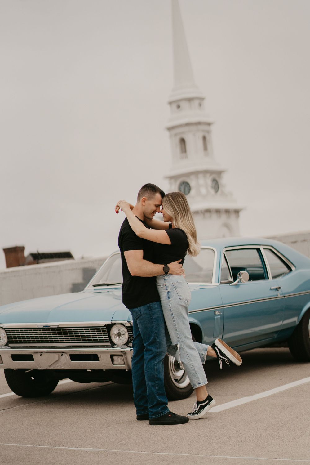 Romantic couple embracing in front of vintage blue car with church steeple in background.