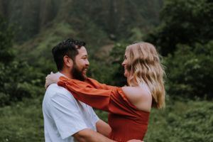 Playful couple embracing in white and rust outfits against green mountain landscape.