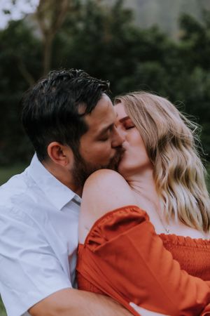 Tender kiss between couple wearing white shirt and rust dress outdoors.