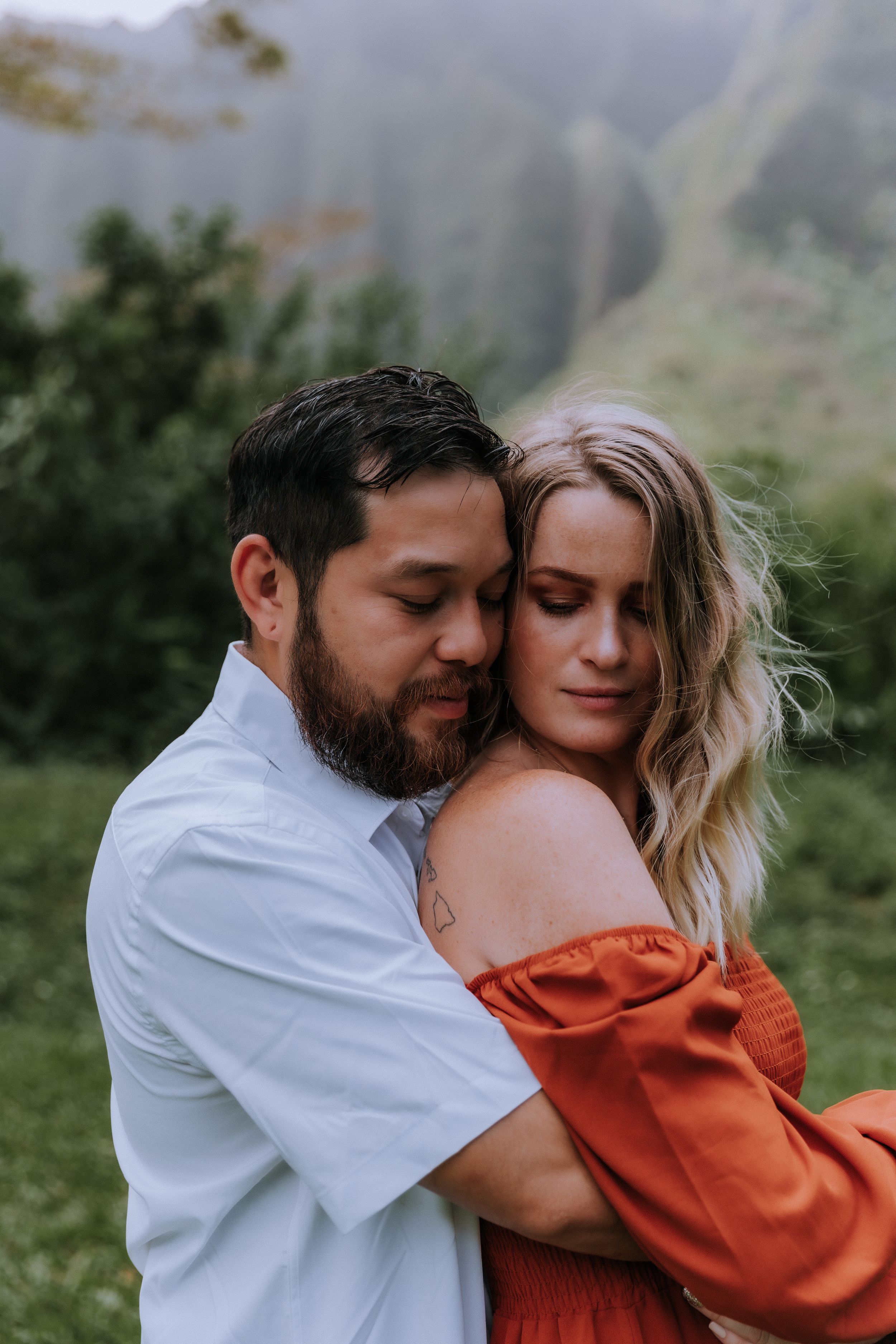 Intimate moment between couple in white shirt and rust dress against mountain backdrop.
