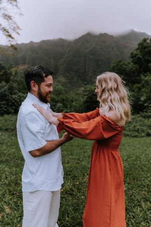 A couple in white and orange outfits playfully interact against a misty mountain backdrop in Hawaii.