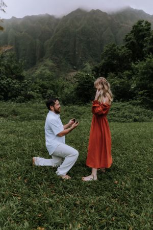 A surprise proposal moment unfolds in a scenic mountain valley with green foliage.