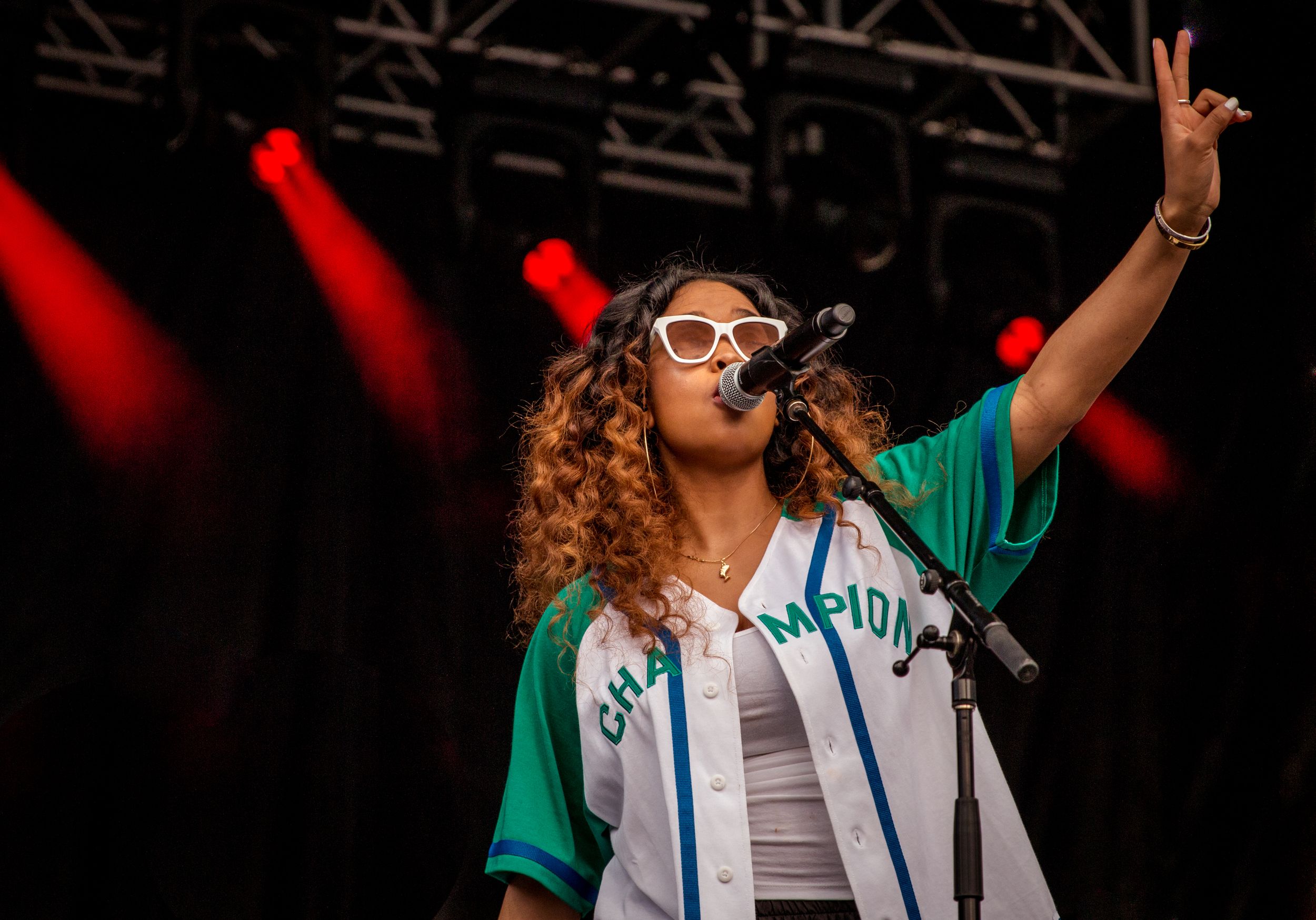 Performer in green jersey and sunglasses singing on stage with arm raised.