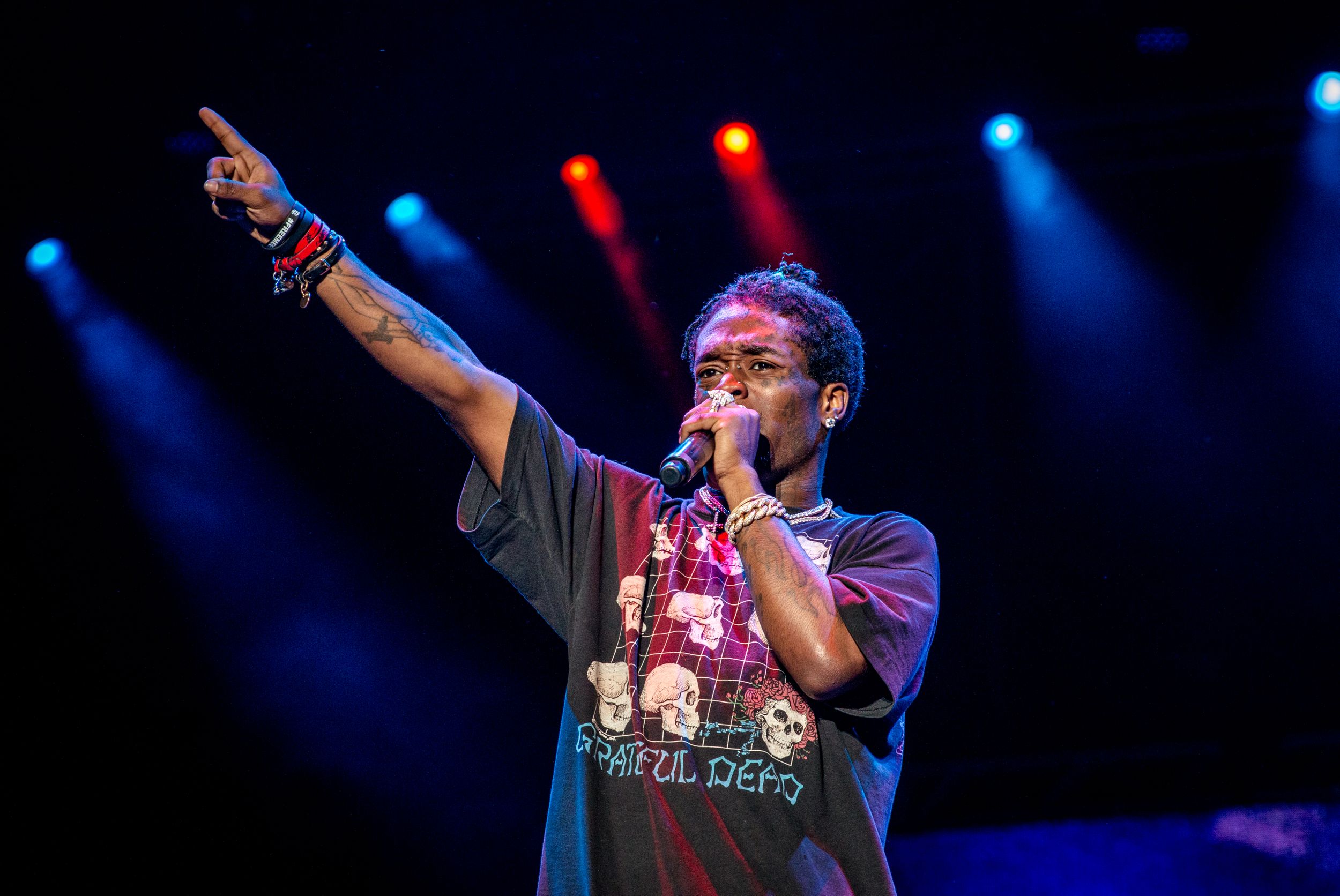 Performer in dark clothing gesturing on stage with colorful lighting and spotlights.