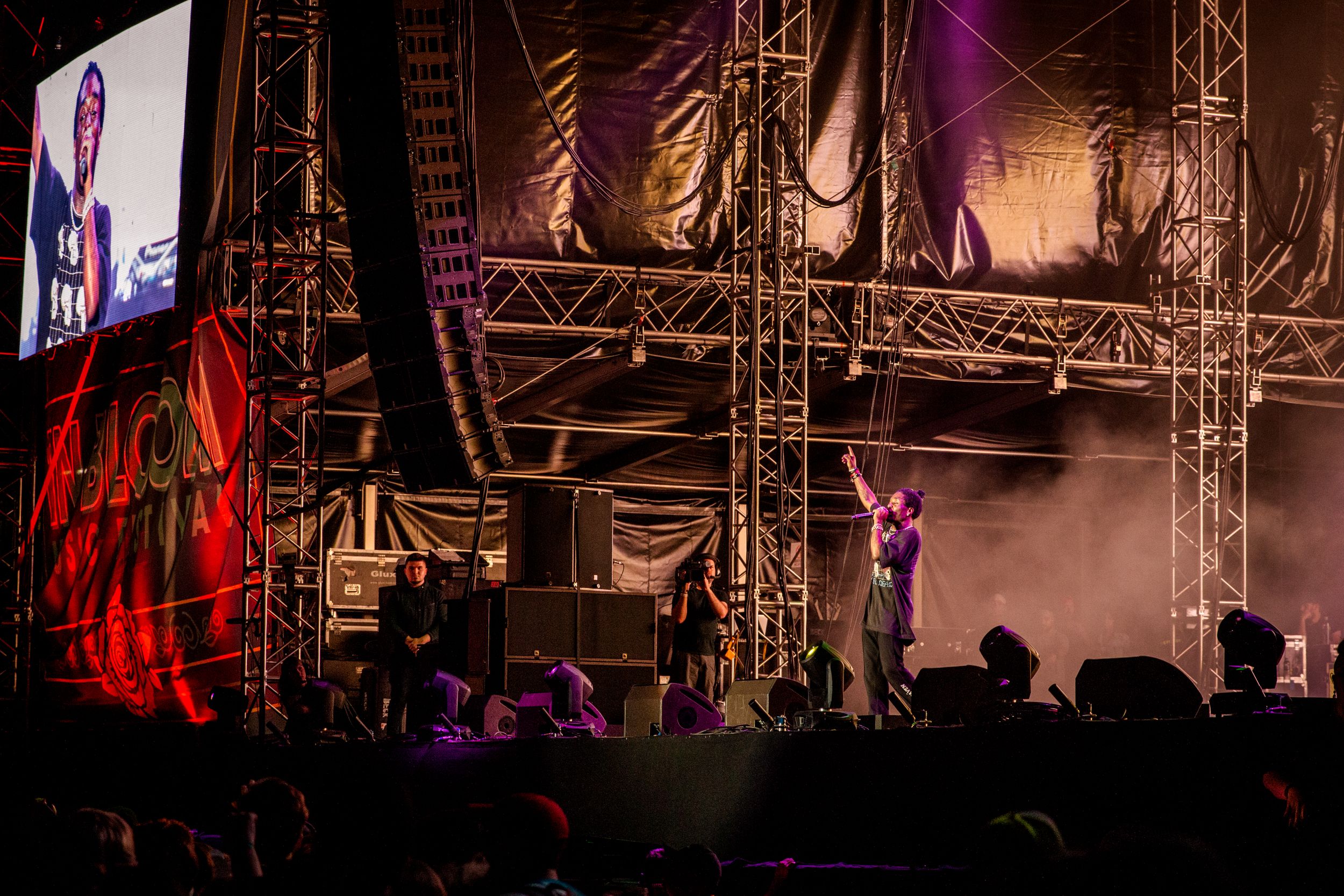 A performer on stage at night with spotlights and a large video screen displaying their image.