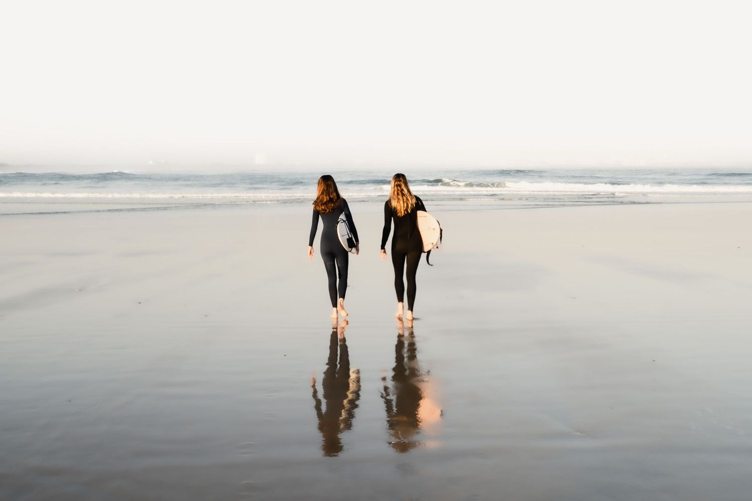 Two surfers walking on wet beach sand at sunrise with their reflections visible.