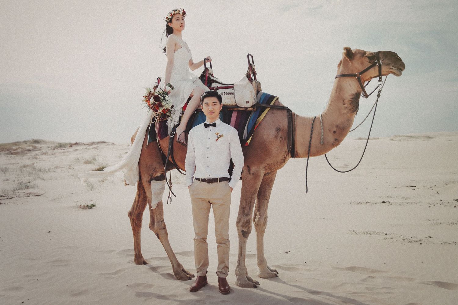 Bride and groom posing with camel on white sand dunes in desert wedding photo.