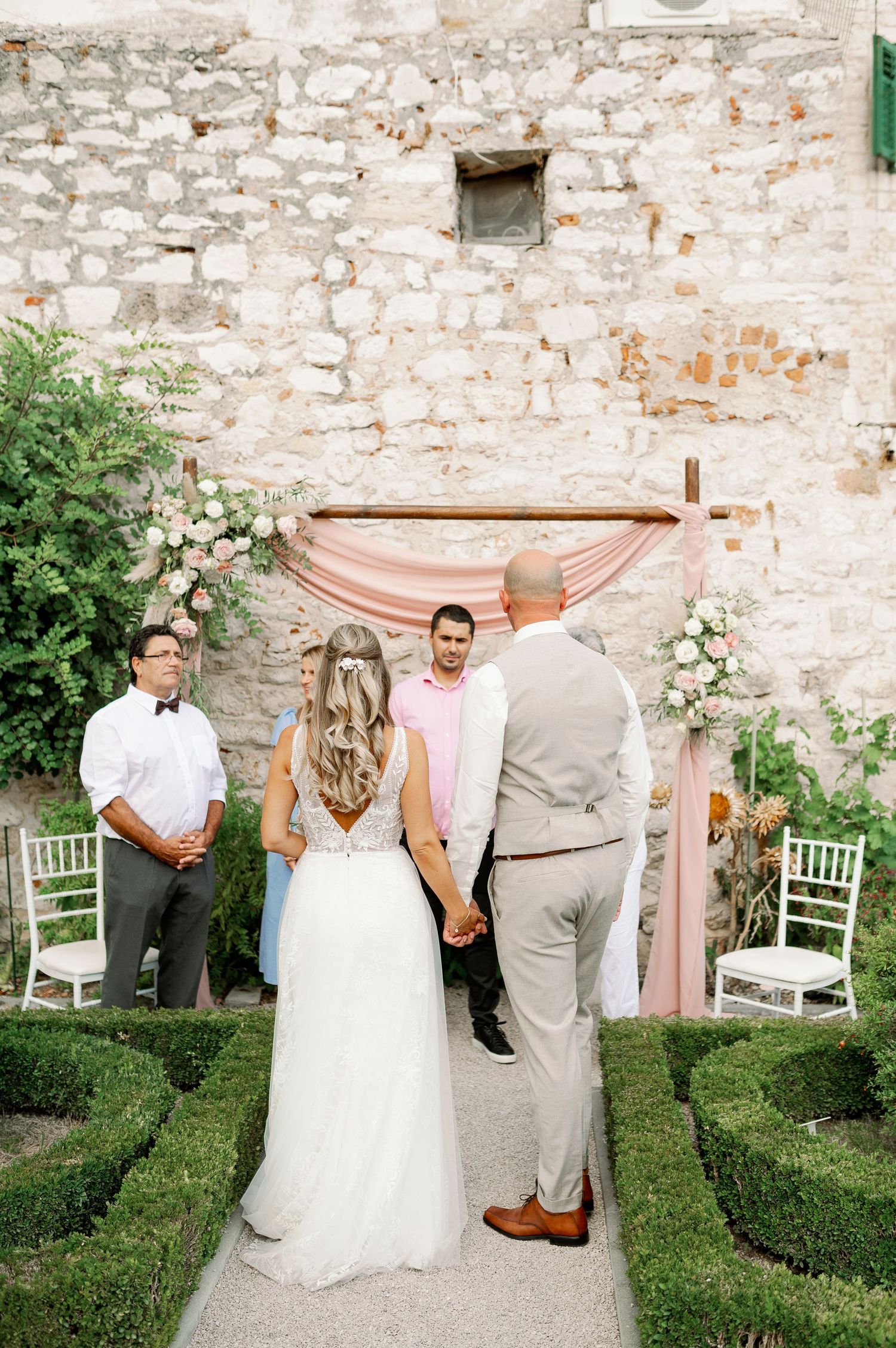 An intimate outdoor wedding ceremony in front of a stone wall with a blush pink fabric arch.