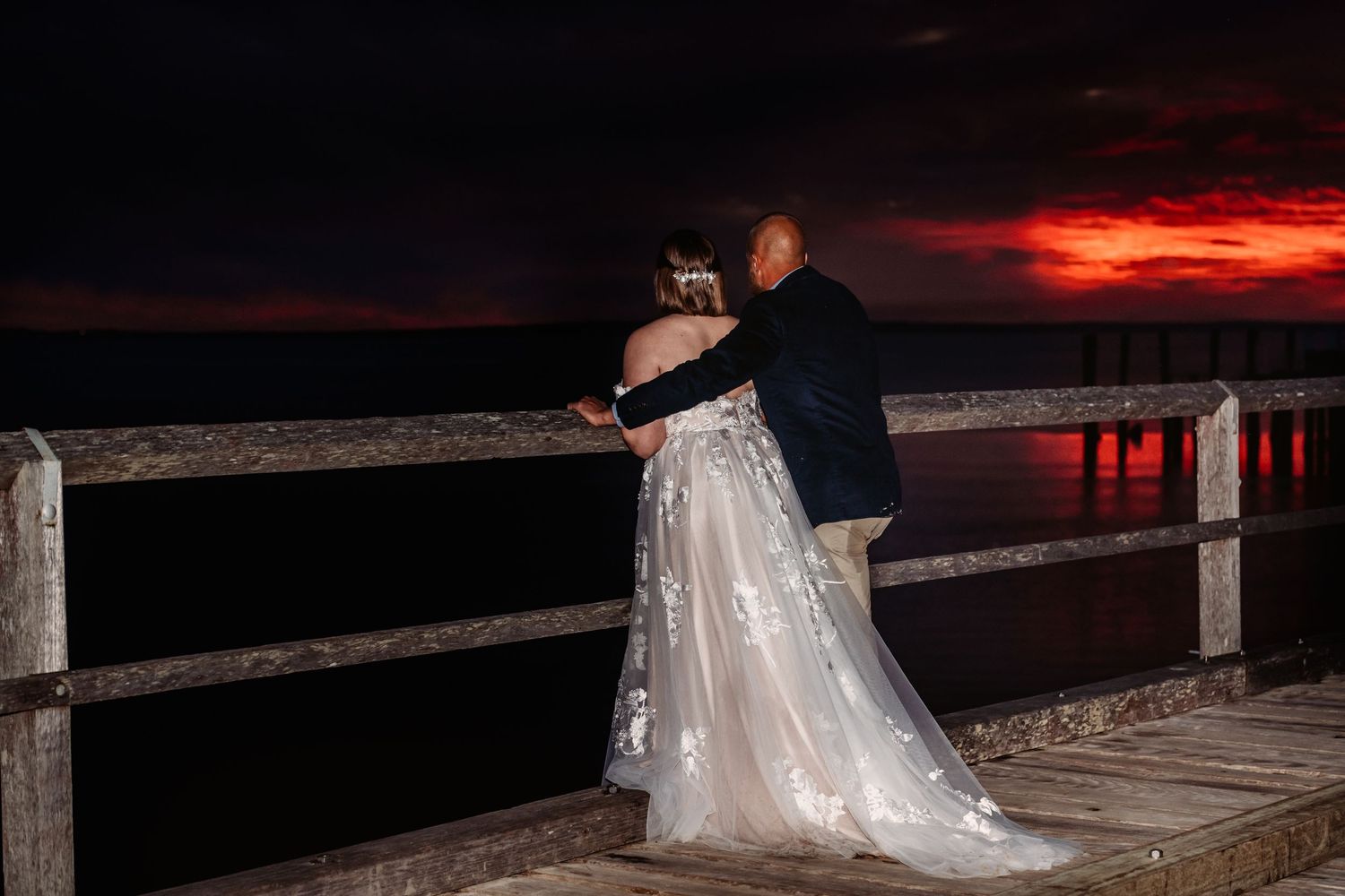 Couple standing at railing watching dramatic red sunset over ocean.