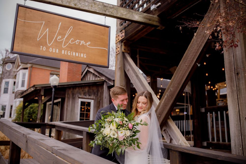 Newlyweds stand beneath rustic welcome sign with elegant floral bouquet.