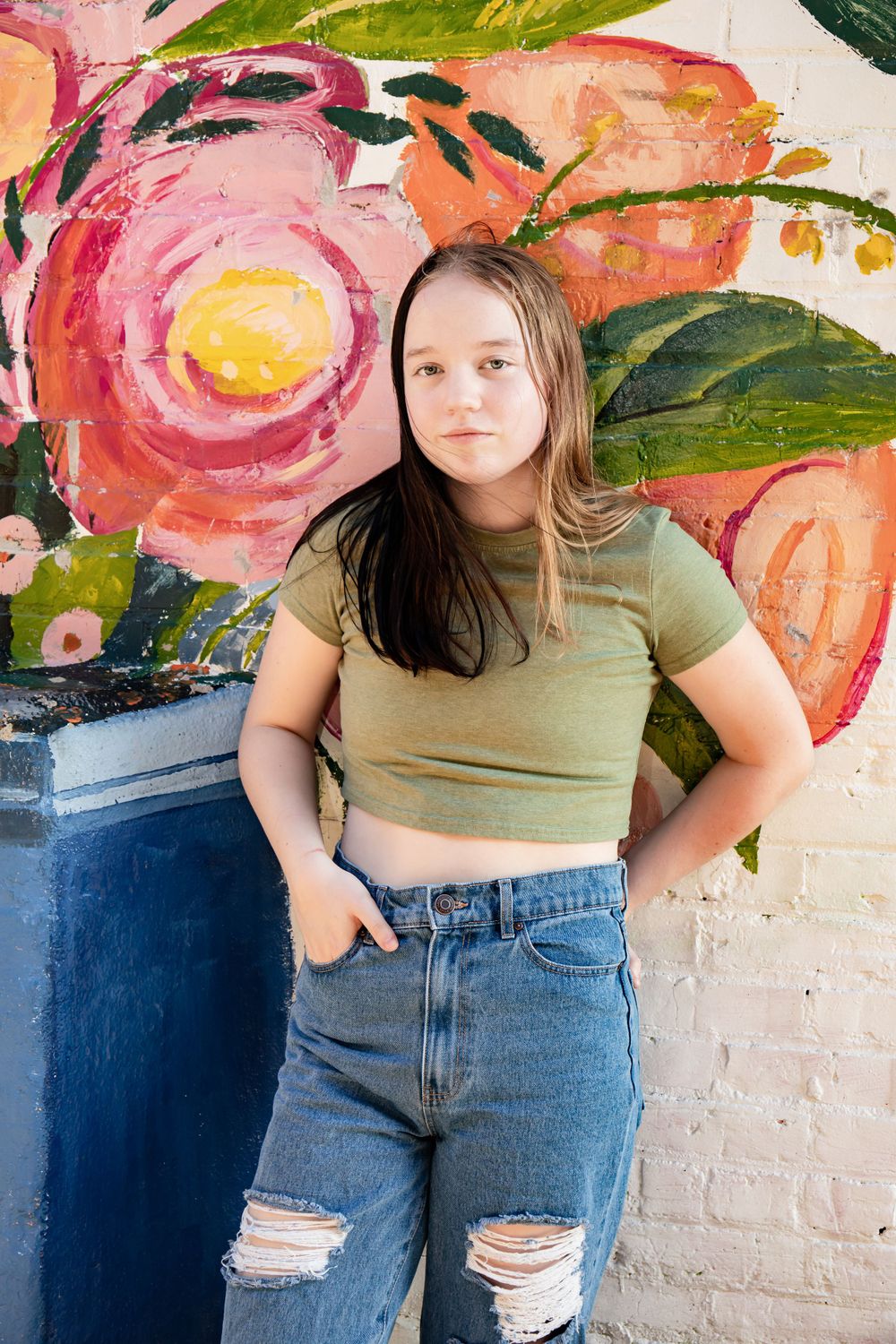 Person in crop top and ripped jeans poses against vibrant floral wall mural.