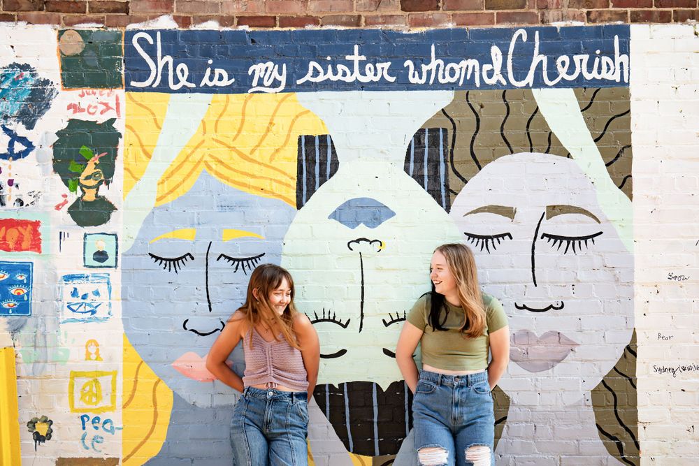 Two people pose against a colorful mural with faces and text on a brick wall.