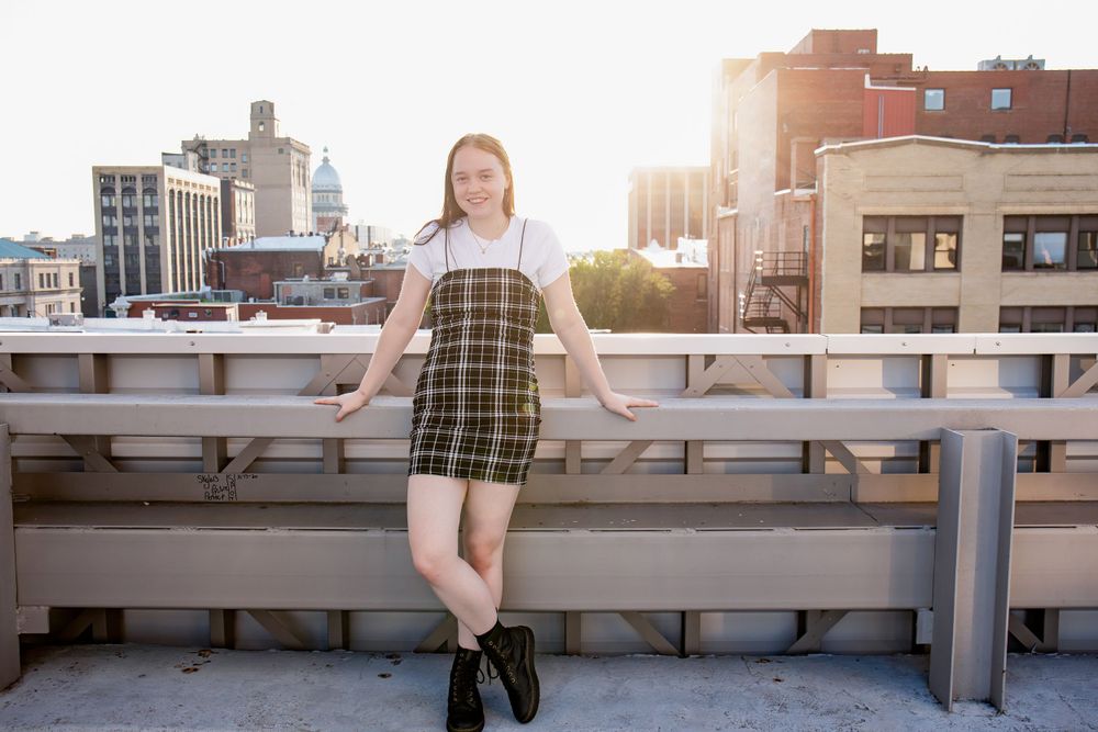 Urban fashion shoot on rooftop with plaid overall dress and combat boots against city skyline.