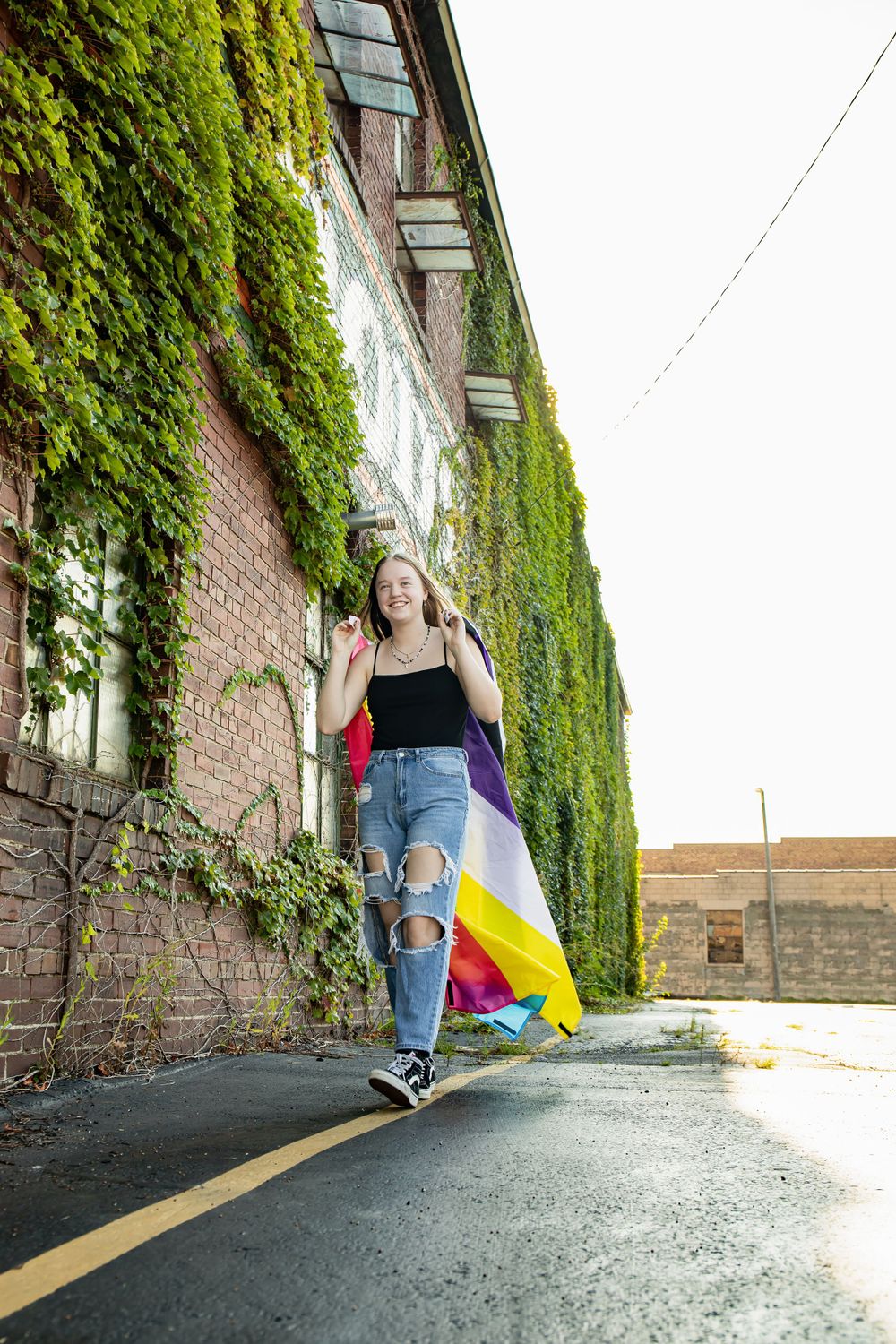 A figure with a pride flag walks along an ivy-covered brick wall in casual attire.