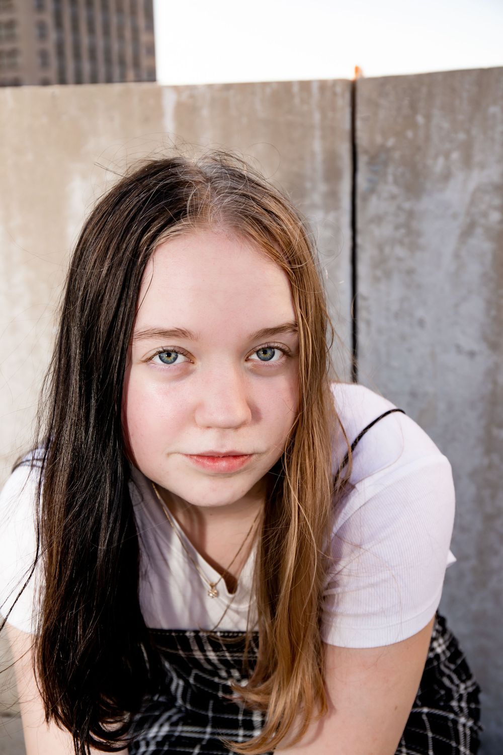 Close-up portrait featuring plaid overall dress and white t-shirt against urban backdrop.