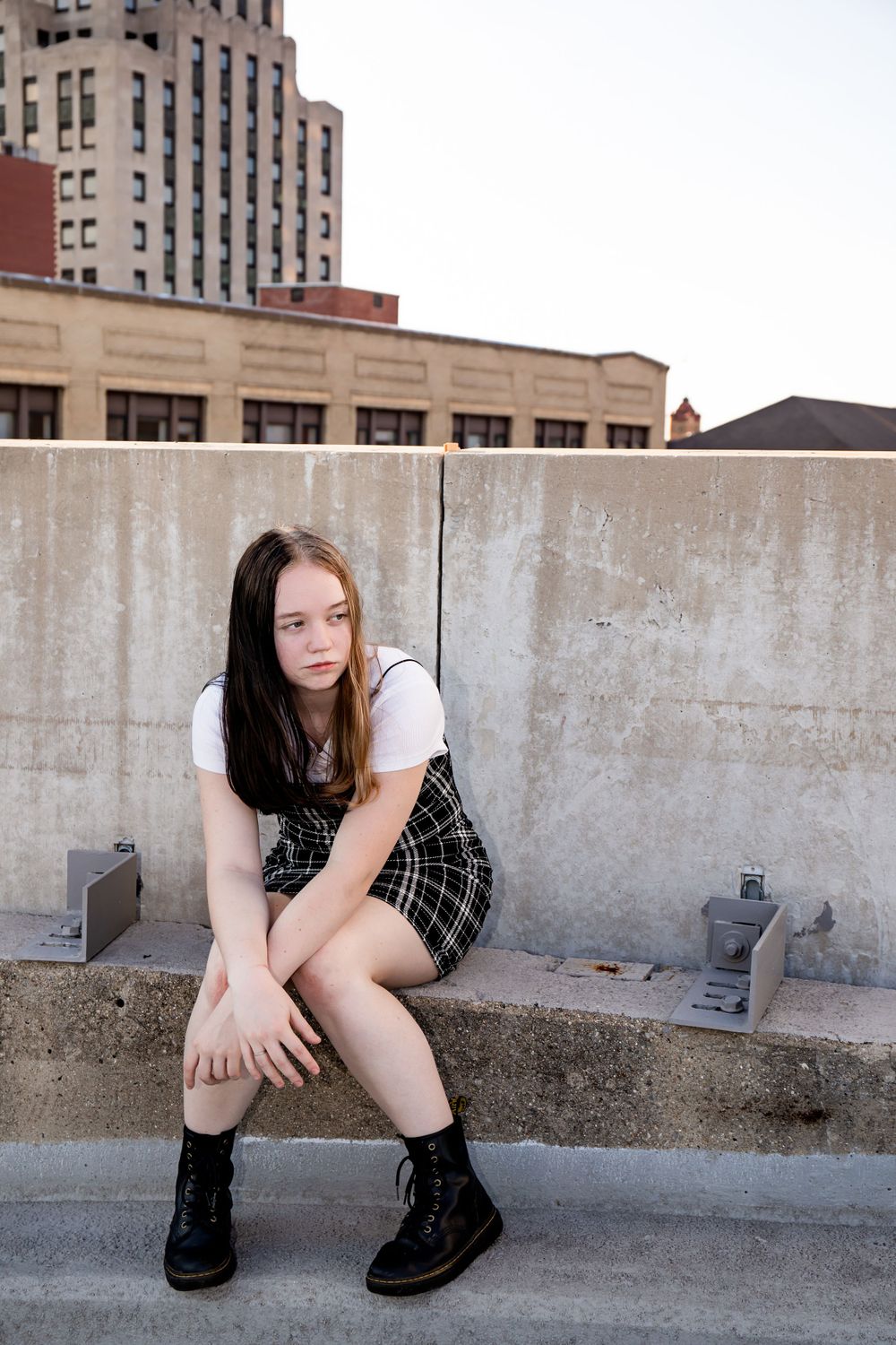 Figure sitting on concrete steps wearing combat boots, a plaid skirt and white top against an urban backdrop.