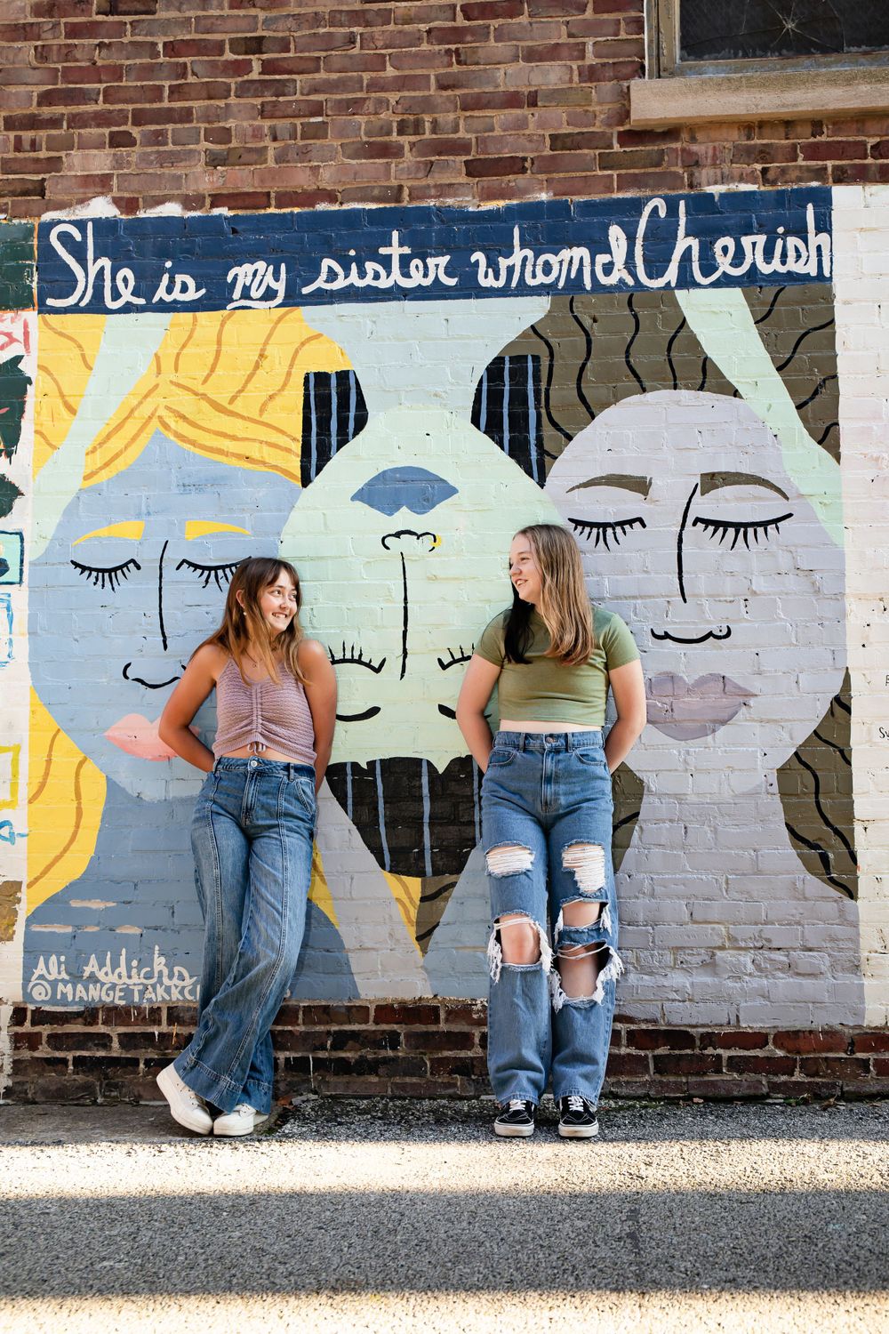 Two people standing against street art mural featuring stylized faces and text.
