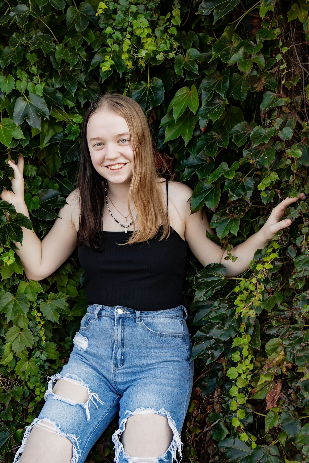 Person in black top and ripped jeans poses against wall of green ivy leaves.
