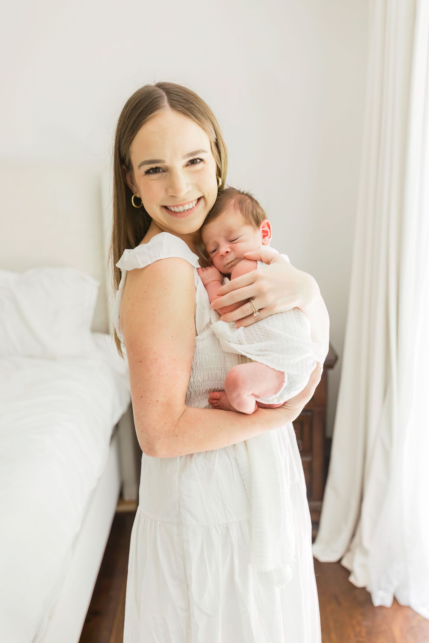 Parent in white dress holding newborn baby against bright interior background.