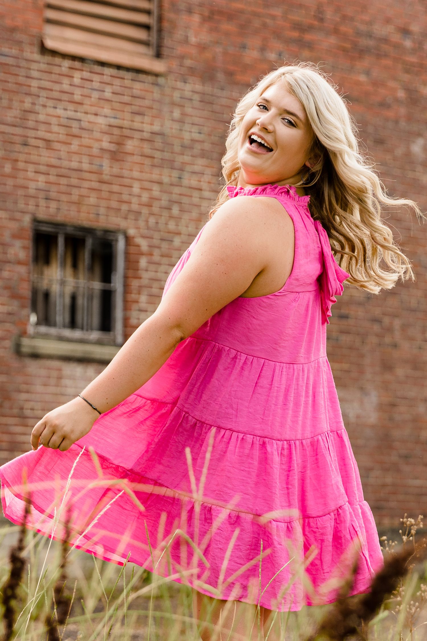 Laughing blonde woman in pink tiered dress twirling near a rustic brick building outdoors.