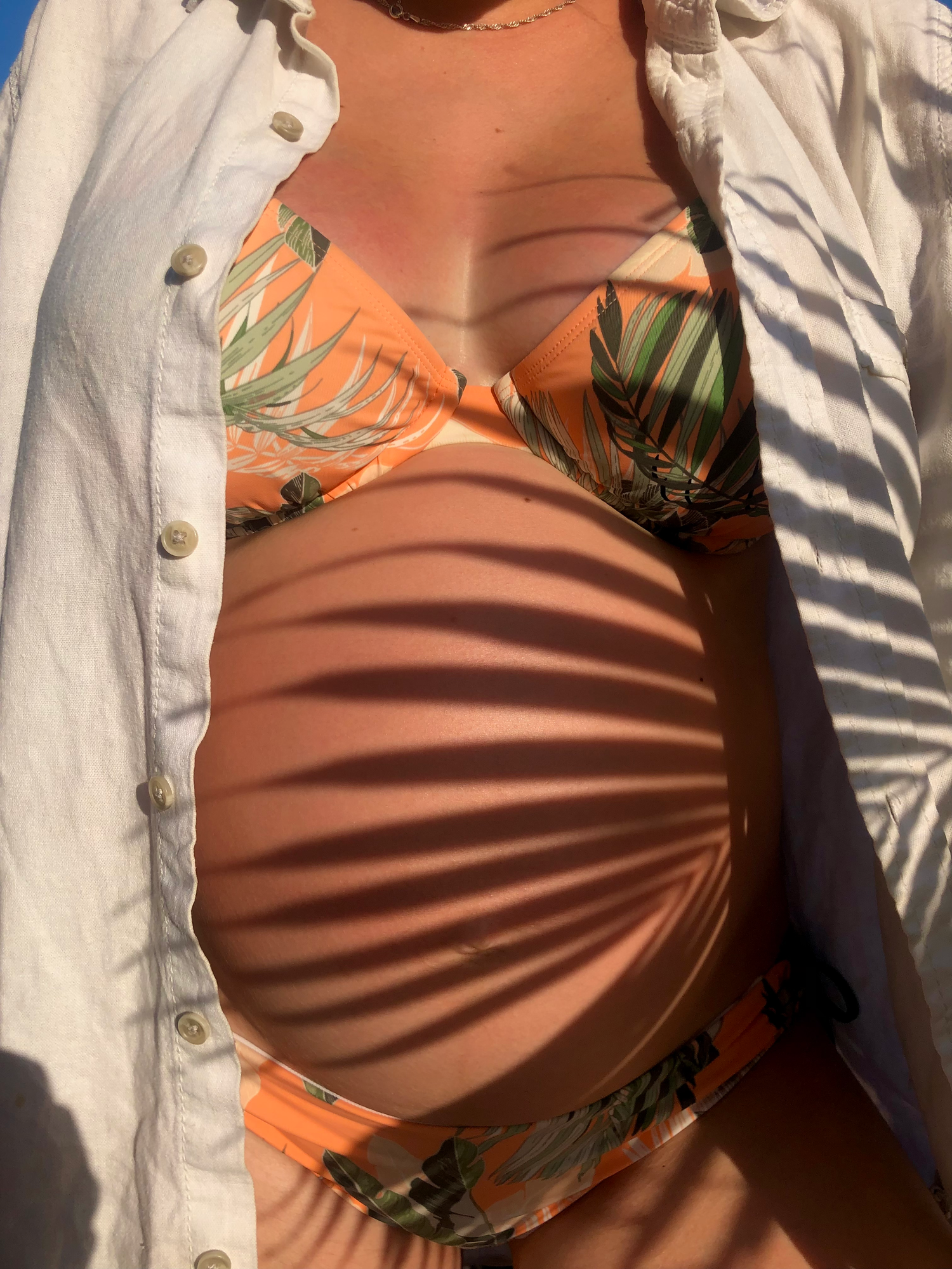 Pregnant woman in orange tropical print bikini and white linen shirt with palm leaf shadows outdoors.