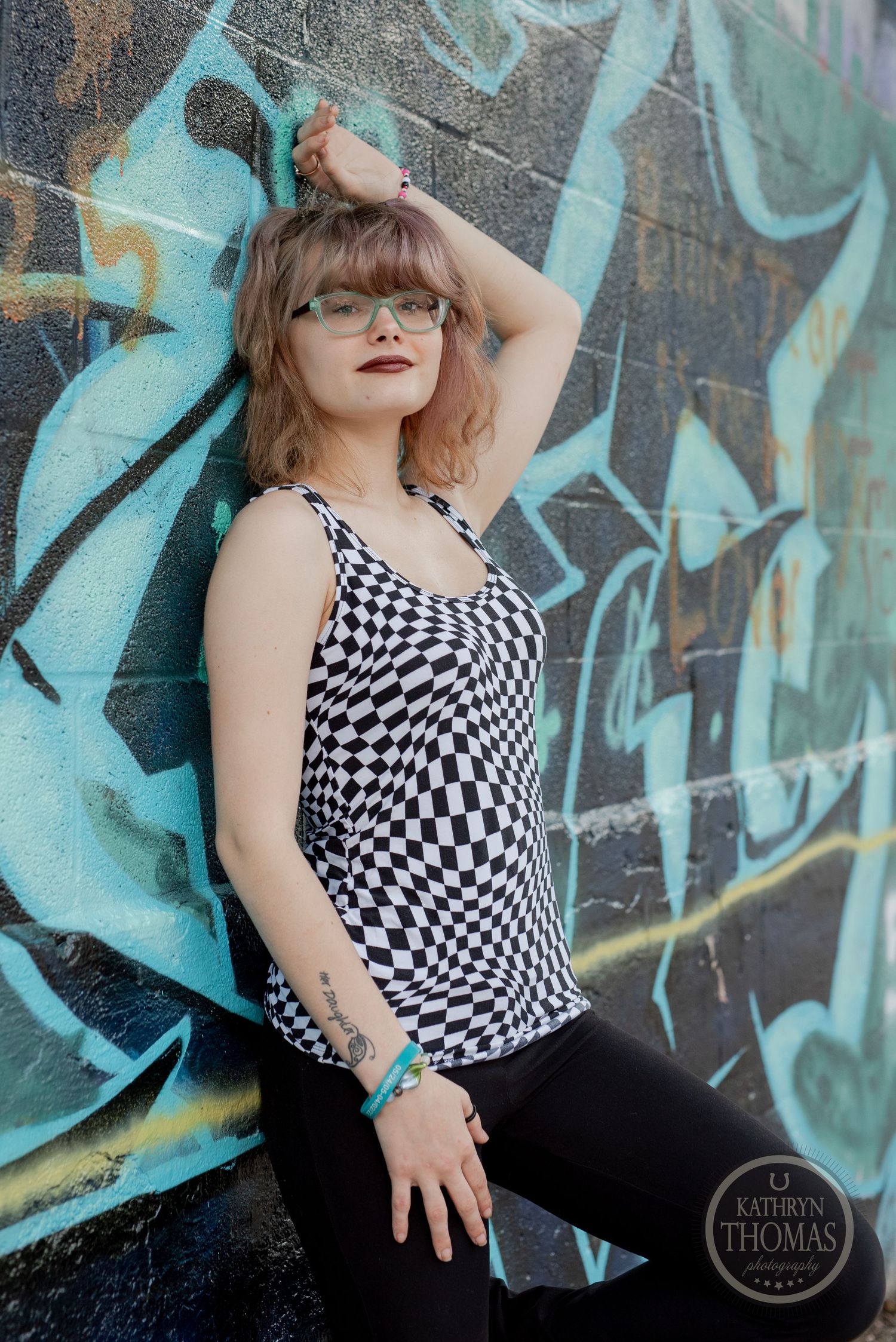 A person poses against turquoise graffiti wall wearing black and white checkered tank top.
