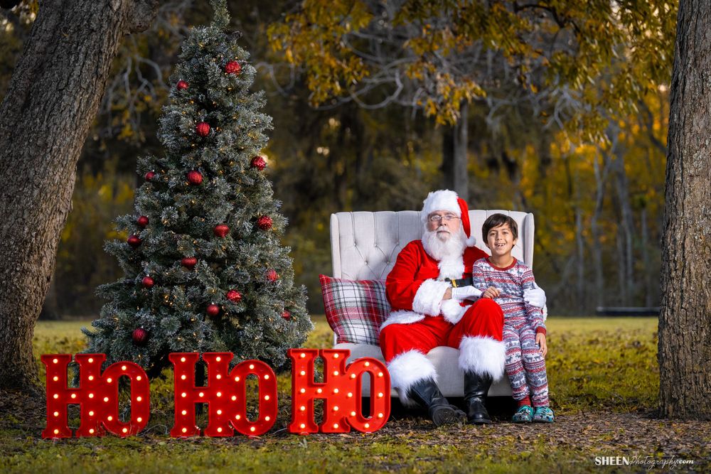 A Christmas photo scene with decorated evergreen tree and wrapped presents against golden fall foliage.